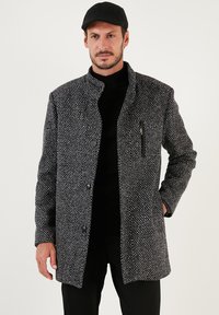 Abrigo texturizado en blanco y negro con un alto cuello, botones en el frente y un bolsillo con cremallera. Se lleva sobre un suéter de cuello alto negro y se acompaña de una gorra.