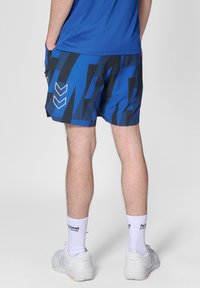 Blå sportshorts med ett geometriskt svart mönster, elastisk midja, sidofickor och logotypdetaljer. Kombinerade med vita strumpor och skor.