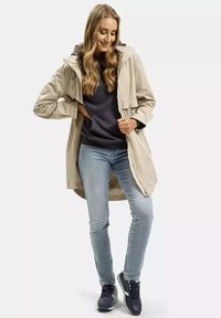 Beżowy lekki płaszcz z kapturem, elastycznym pasem, noszony na ciemnej bluzie z kapturem, w połączeniu z niebieskimi dżinsami i czarnymi trampkami. Gładka tekstura, casualowy design.