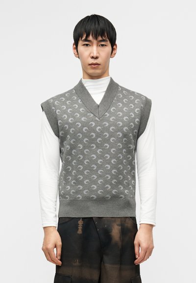 Γκρι πλεκτό γιλέκο χωρίς μανίκια με διακριτικό σχέδιο ημισελήνου, σχέδιο με λαιμόκοψη σε σχήμα V και απαλή υφή, φορεμένο πάνω από λευκή μακριμάνικη μπλούζα.