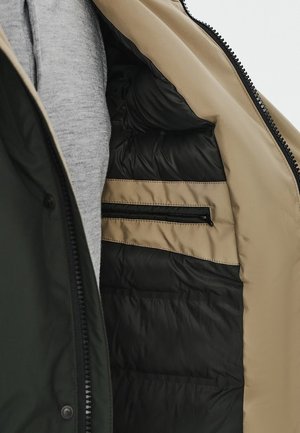 Zielona i beżowa kurtka wewnątrz ma pikowaną czarną podszewkę, szary materiał z tyłu oraz jedną kieszeń na zamek błyskawiczny w beżowej części.