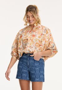 Blouse florale à manches bouffantes dans des couleurs pastel et un plastron à demi-boutons, accompagnée de shorts en denim bleu avec un motif décoratif.