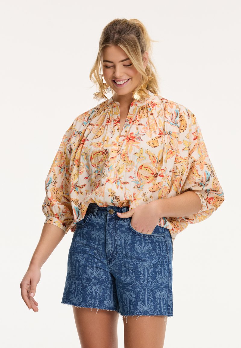 Blouse florale à manches bouffantes dans des couleurs pastel et un plastron à demi-boutons, accompagnée de shorts en denim bleu avec un motif décoratif.