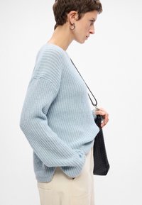 Jasnoniebieski sweter robiony na drutach o luźnym kroju, o żebrowanej teksturze i obniżonych ramionach, w połączeniu z kremowymi spodniami i czarną torebką o fakturze.