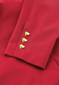Rode stof met een gladde textuur heeft drie gouden hartvormige knopen nabij de manchetten, met subtiele stiksels die het ontwerp accentueren.
