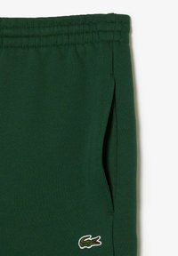 Pantalones de chándal verdes de algodón con cinturilla elástica, bolsillos laterales y un pequeño logo bordado cerca del dobladillo. Textura suave.