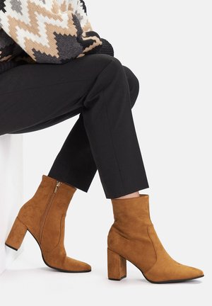 Brązowe zamszowe botki za kostkę z noskiem w szpic i obcasem typu blok. Posiadają boczny zamek oraz gładką fakturę, zestawione z czarnymi spodniami.