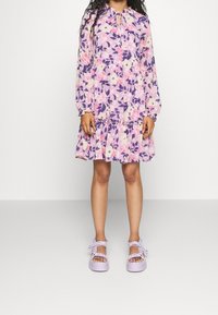 Květinové šaty s dlouhými rukávy v odstínech fialové a růžové, s nabíraným pasem a volánkovým lemem. Doplněné levandulovými sandály.