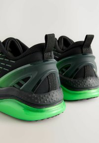 Chaussures de sport noires avec une tige respirante, une semelle translucide verte, des accents texturés et une languette au talon pour un enfilage plus facile.