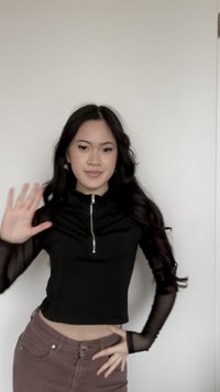 Mlada ženska z dolgimi temnimi lasmi, oblečena v črno mrežasto majico z dolgimi rokavi in rjave hlače, ki se smehlja in maha z desno roko.