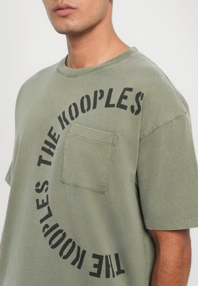 T-shirt de algodão verde-azeitona com decote redondo, mangas curtas, bolso de remendo frontal e design de logotipo curvo em preto. Textura suave.