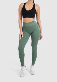 Zelené proužkované legíny s vysokým pasem v kombinaci s černým sportovním podprsenkou. Modelka má na sobě bílé tenisky. Jednoduché světlé pozadí.