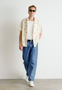 Majica s kratkimi rokavi, svetle barve z zelenimi vzorci, bela majica, ohlapne modre jeans hlače in bele športne copate; nosijo se sončna očala.