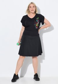 Zwarte sportrok gecombineerd met een zwart V-hals T-shirt met korte mouwen en een kleurrijke "SOL"-grafiek. Model houdt een groene dumbbell vast.