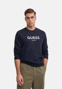 Felpa blu navy con collo tondo e polsini a coste, con il logo "GUESS EST. 1981" stampato in bianco sul petto. Tessuto in cotone misto tessurato.