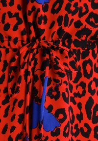 Tela roja con un patrón de print de leopardo negro. Se puede ver un acento azul, atado en la cintura con un cinturón de tejido a juego, que presenta una textura suave.