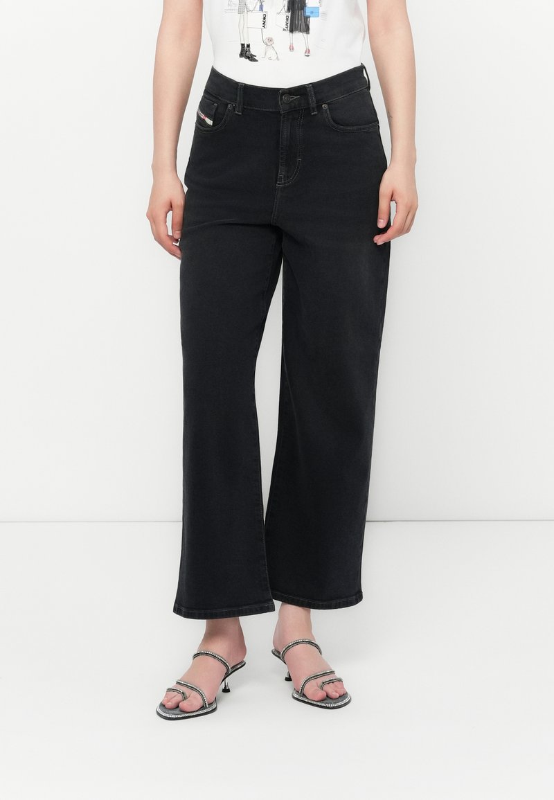 Jean noir à jambes larges en denim, avec une taille haute, cinq poches et des coutures tonal subtils. Associé à des sandales à lanières minimalistes.
