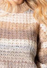 Pull en maille épaisse avec des rayures horizontales dans des tons beige, crème, marron clair et gris, porté par une personne aux cheveux blonds.