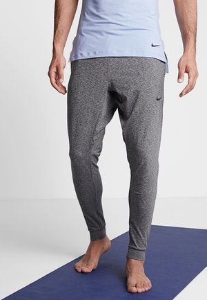 Homme portant un pantalon de jogging Nike gris et un t-shirt Nike bleu clair, debout pieds nus sur un tapis de yoga bleu avec un fond uni.