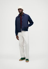 Tumši zils žakete ar pogu aizdari pār bordo krāsas polo krekli, saskaņots ar gaišas krāsas biksēm un zaļām sporta kurpēm. Modelis valkā krēmkrāsas cepuri.