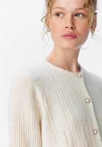 Cremfarbener Strickcardigan mit strukturierten Streifen, rundem Halsauschnitt und drei perlenähnlichen Knöpfen. Weiches, gemütliches Material mit gerippten Ärmeln.