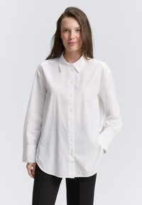 Camicia bianca con colletto classico, maniche lunghe e taglio dritto. Realizzata in un tessuto leggero e liscio con una sottile trama.