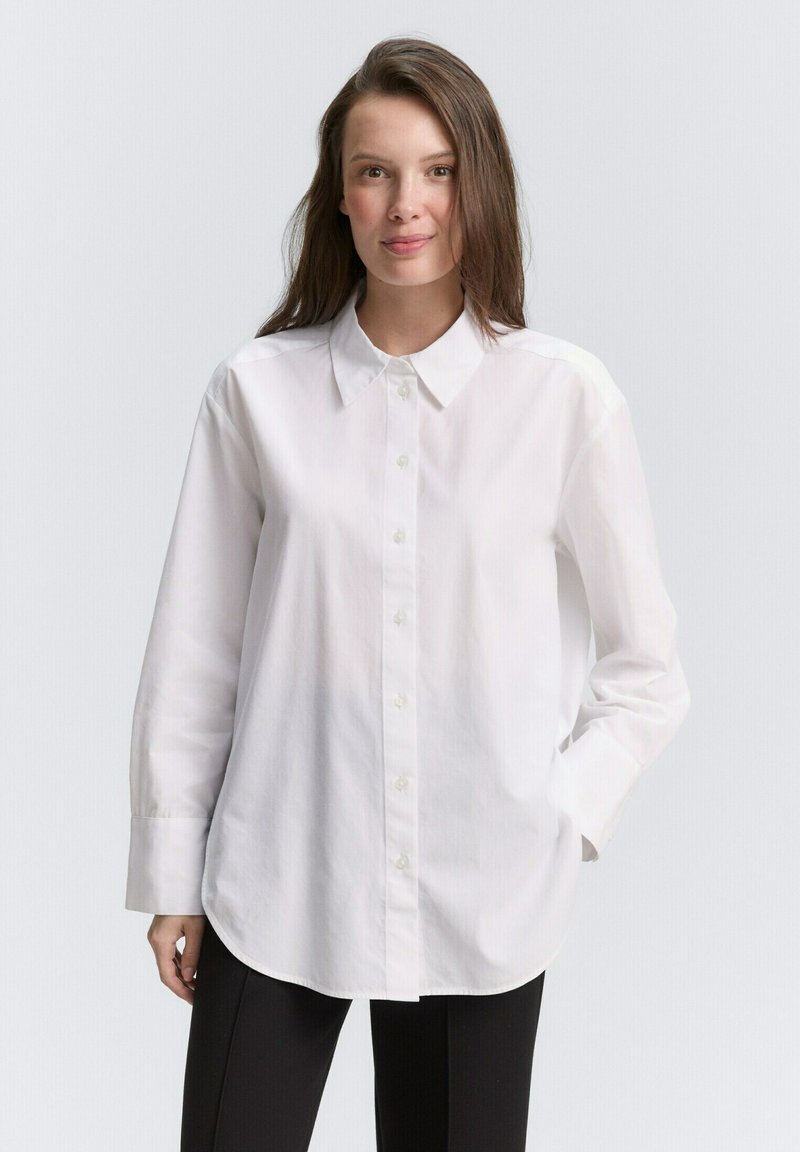 Camicia bianca con colletto classico, maniche lunghe e taglio dritto. Realizzata in un tessuto leggero e liscio con una sottile trama.