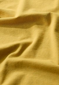 Tekstili kangas, pehme puuvill, sinep kollane värv, sile tekstuur kergete kortsude ja voldidega, ühtlane kudumise muster kogu pinna ulatuses.