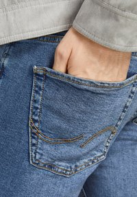 Jeans din denim cu o nuanță albastră, cu cusături vizibile de culoare aurie, o buză din spate și o mână introdusă în buzunar.