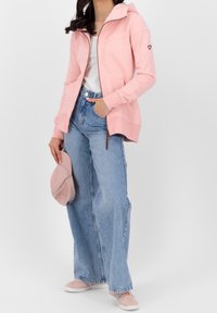 Donna che indossa una felpa rosa chiaro con zip, una maglietta bianca, jeans a gamba larga blu e sneaker rosa, mentre tiene un cappellino rosa in una mano.