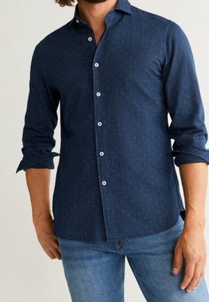 Chemise boutonnée bleu marine avec un motif subtil, manches longues retroussées, en tissu tissé ; elle présente des boutons blancs et un col structuré.