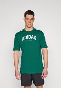 Zöld, rövid ujjú póló fehér "ADIDAS" logóval és csíkokkal a vállakon. Simább anyagból készült, kerek nyakú dizájnnal.