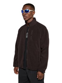 Homme portant une veste polaire marron foncé zippée et un pantalon noir, accessoirisé de lunettes de soleil bleues réfléchissantes avec une monture blanche et une petite boucle d'oreille en forme d'anneau.