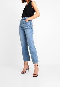 Jean taille haute bleu clair avec une coupe droite et un ourlet brut ; associé à un haut noir sans manches et des sandales à talons hauts noires à brides.