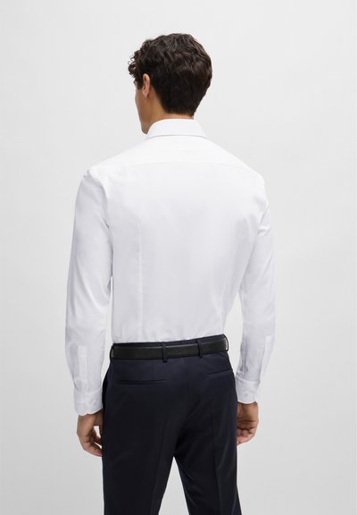 Chemise blanche à bouton, à manches longues, à la texture lisse et à la coupe ajustée, associée à un pantalon foncé et une ceinture noire.