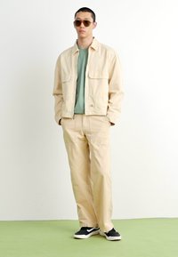 Beige canvas jacket and matching pants, layered over a light green shirt. Sunglasses and black sneakers complete the outfit. Smooth texture.