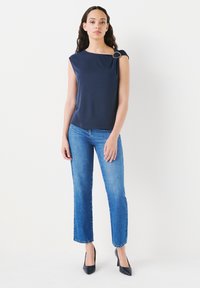 Navy sleeveless top with a gold accent on the shoulder, paired with light blue straight-leg jeans and black pointed-toe heels.