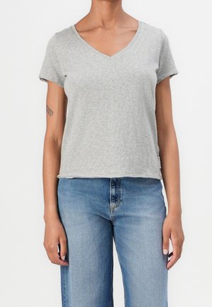 Vrouw draagt een lichtgrijs T-shirt met V-hals en korte mouwen en een blauwe spijkerbroek, staand tegen een witte achtergrond, handen ontspannen langs het lichaam.