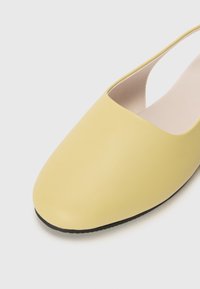 Geltoni slingback bateliai su lygiu sintetiniu paviršiumi, apvalia nosimi ir juoda guminėmis padais. Turintys minimalų dizainą ir lengvai užsidedančią atvartą.