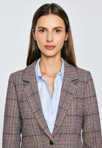 Kostkovaný blazer s kombinací béžových, hnědých a modrých tónů, strukturovaný design, široké klopy a jedno černé zapínání na knoflík.