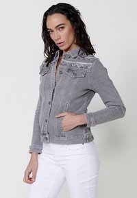Sivina denim jakna z okrasnim šivom ob ramenih, s preklopom na gumb in sprednjima žepoma. Usklajena z belimi kavbojkami.