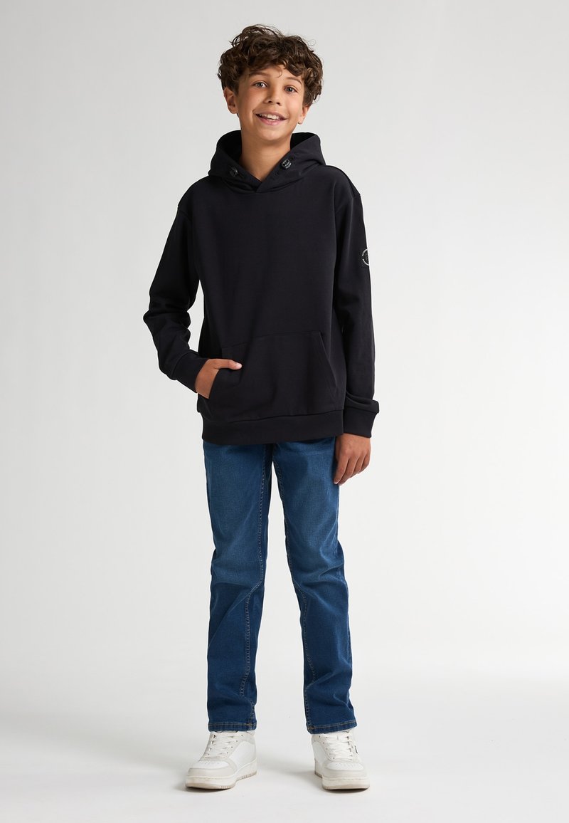 Sudadera con capucha negra, bolsillo frontal y logo en la manga, combinada con jeans azules y zapatillas blancas. Textura suave, corte relajado.