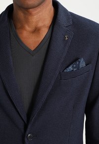 Granatowa teksturowana marynarka na jeden guzik, z poszetką w niebieski wzór paisley, pod spodem ciemnoszara koszulka z dekoltem w serek.