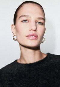Suéter negro de felpa con un escote redondeado, con aretes de aro dorados. Piel clara y cabello peinado hacia atrás, con maquillaje sutil y un fondo neutral.