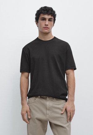 Jonge man met krullend donker haar, gekleed in een effen zwart T-shirt en beige broek, staand tegen een witte achtergrond.