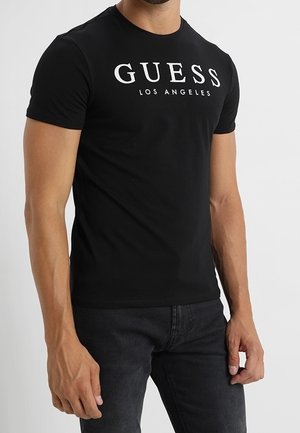 Mężczyzna noszący dopasowany czarny t-shirt z krótkim rękawem z napisem „GUESS LOS ANGELES” oraz ciemne jeansy na jednolitym jasnym tle.