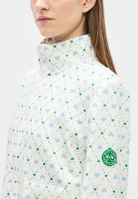 Λευκό μπουφάν με πράσινο και μπλε σχέδιο καρδιάς, ψηλό γιακά, φερμουάρ στο κλείσιμο και σήμα λογότυπου στο μανίκι. Μαλακό ύφασμα, εφαρμοστό σχέδιο.