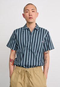 Camisa de mangas curtas em listras verticais azul-marinho e azul-claro, apresentando um colarinho clássico, fecho com botões e um bolso no lado esquerdo do peito.