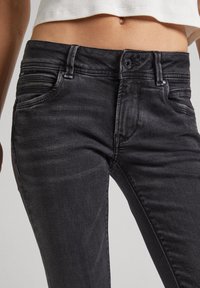 Jeans de mezclilla negra con corte ajustado, que cuentan con un bolsillo con cremallera, costuras visibles y un cierre de botón en la cintura.