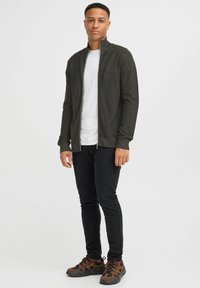 Cardigan zippé gris foncé avec un motif de tricot texturé, porté par-dessus un t-shirt blanc, associé à un pantalon noir et des baskets marron avec des accents.